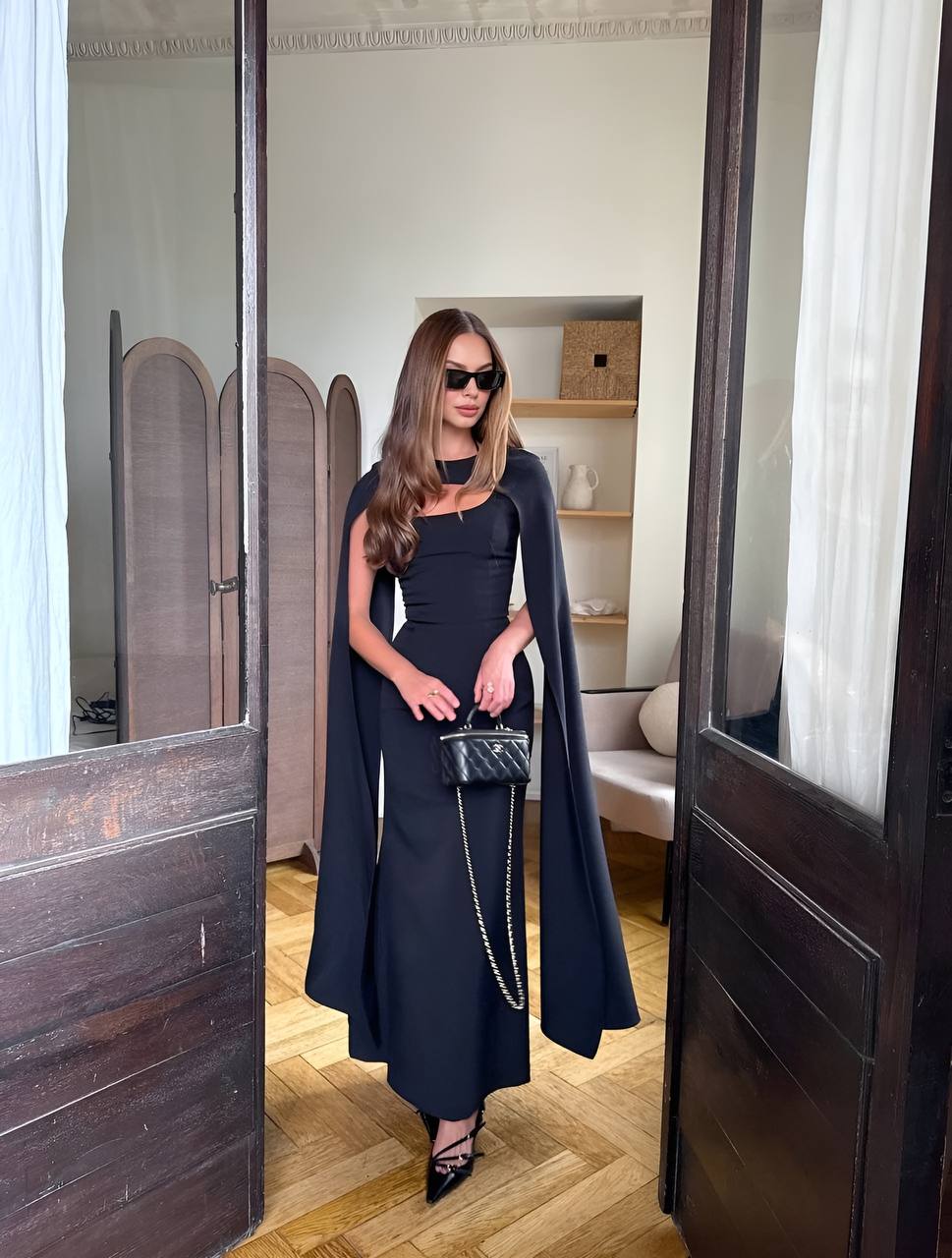Woman in a black dress standing in a room with wooden floor and white walls.