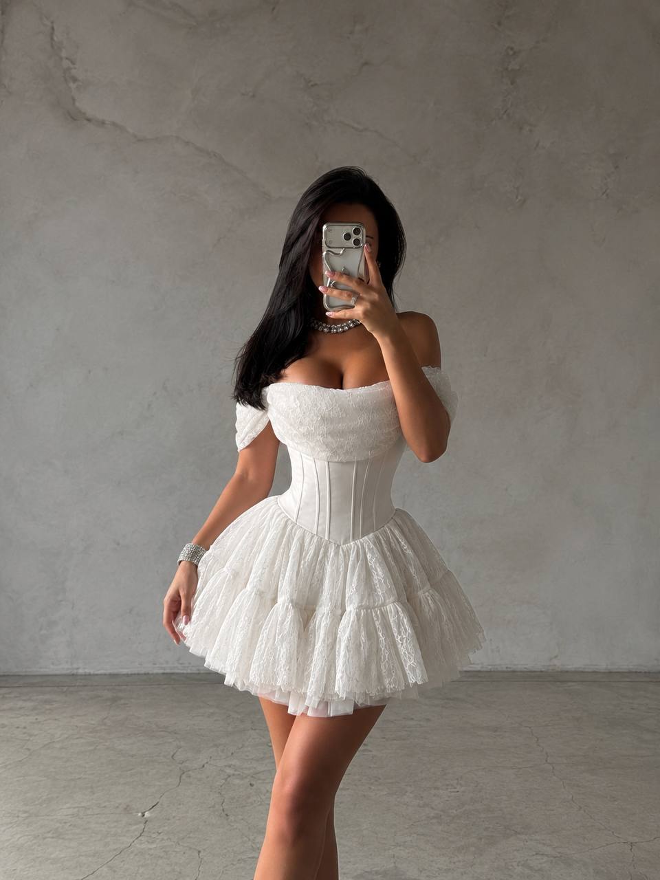 A woman takes a mirror selfie wearing a white off-the-shoulder mini dress with a structured corset-style bodice and a layered ruffled tulle skirt, standing against a textured gray concrete wall.