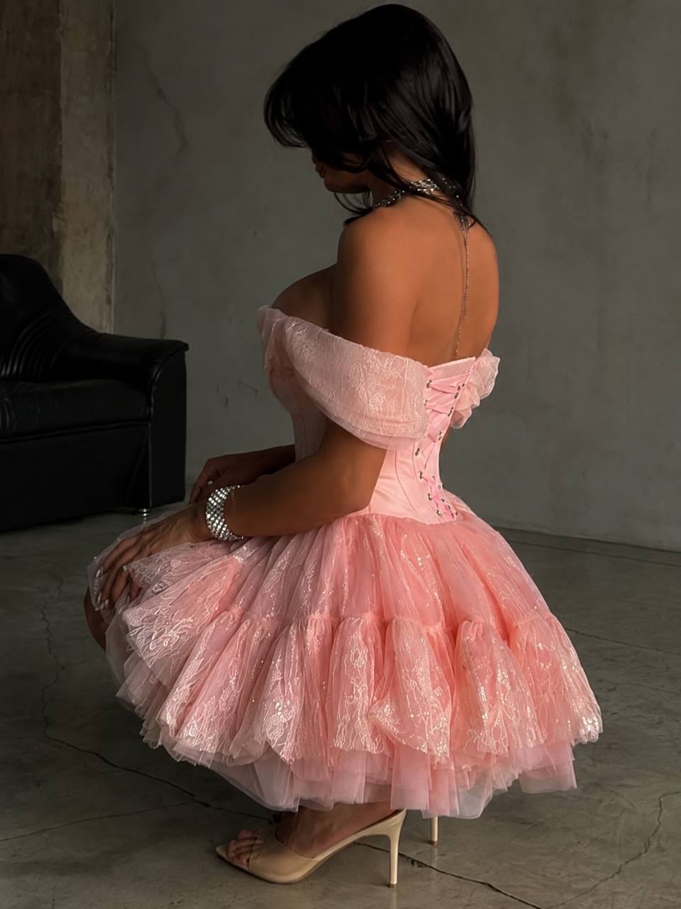 A woman wearing a light pink off-the-shoulder tulle dress with a ruffled corset-style bodice and layered skirt, posed in a dimly lit room with a concrete wall and black chair in the background.