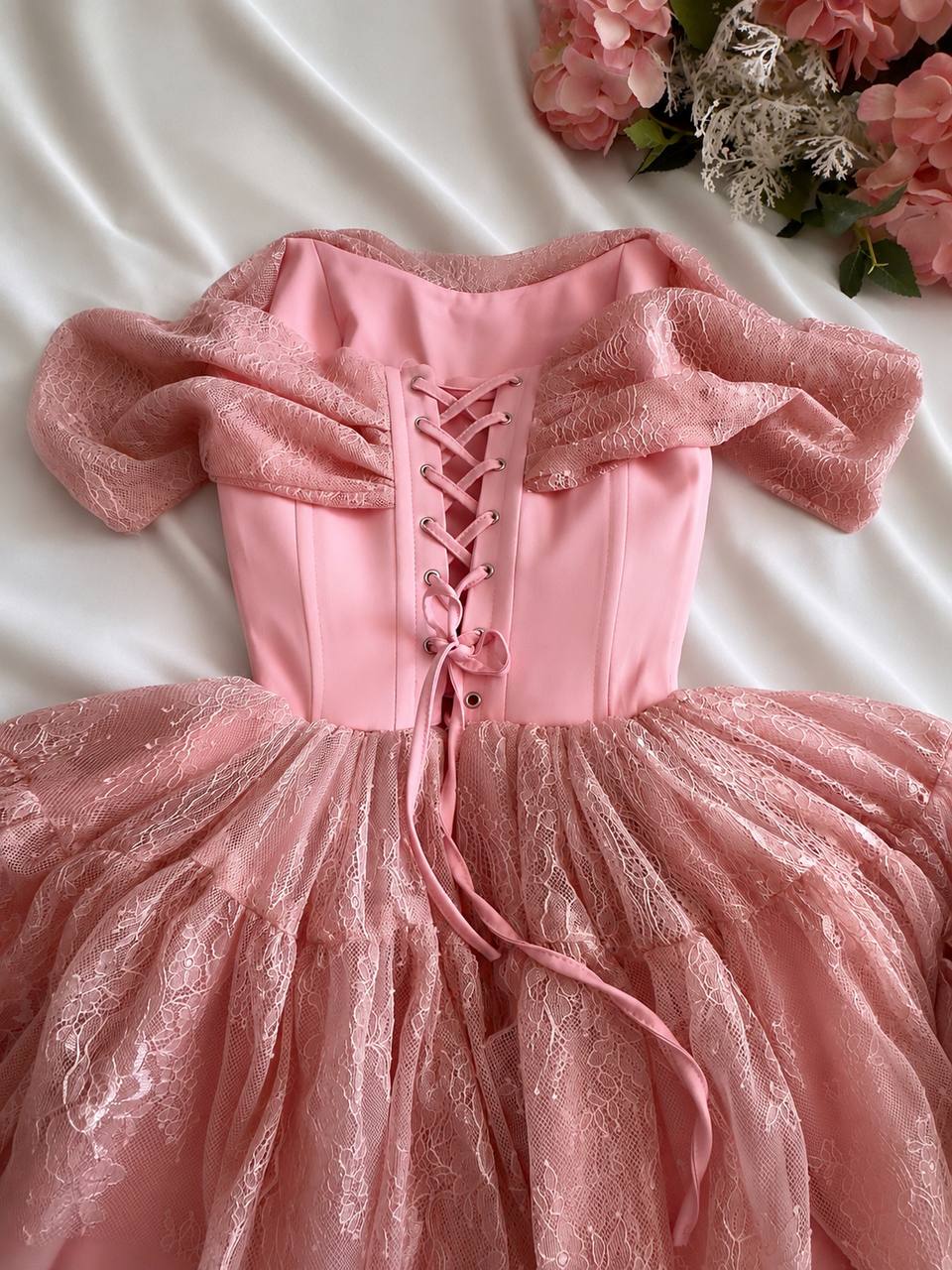 A pink off-the-shoulder dress with a lace-up corset back, puffy sleeves, and a voluminous tulle skirt, laid flat on a white satin sheet with pink flowers in the corner.