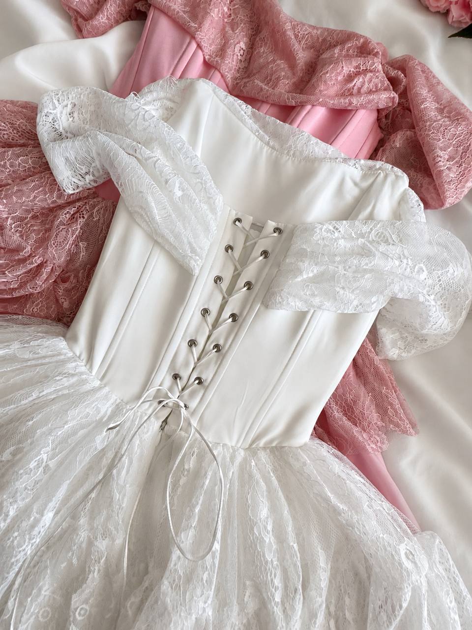 A close-up of a white lace and satin dress laid flat on a white fabric surface, showing the back of the bodice with lace-up detailing and sheer puff sleeves. Pink lace fabric is partially visible underneath.