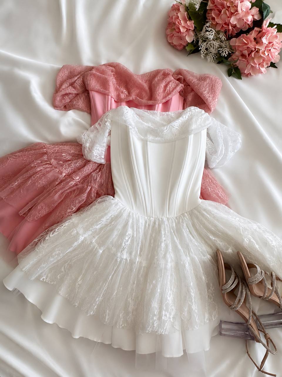 A styled flat-lay of two short dresses on white satin fabric, featuring a white lace corset-style dress in the foreground and a pink velvet and lace dress behind it, with pink flowers and beige strappy shoes as props.