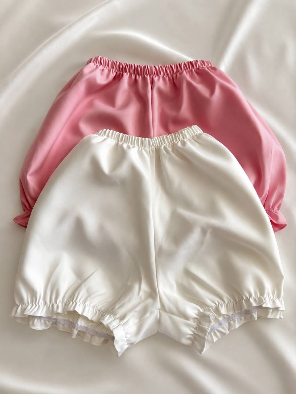 Two satin-like bloomers or shorts in pink and off-white, arranged overlapping on a soft white fabric background.