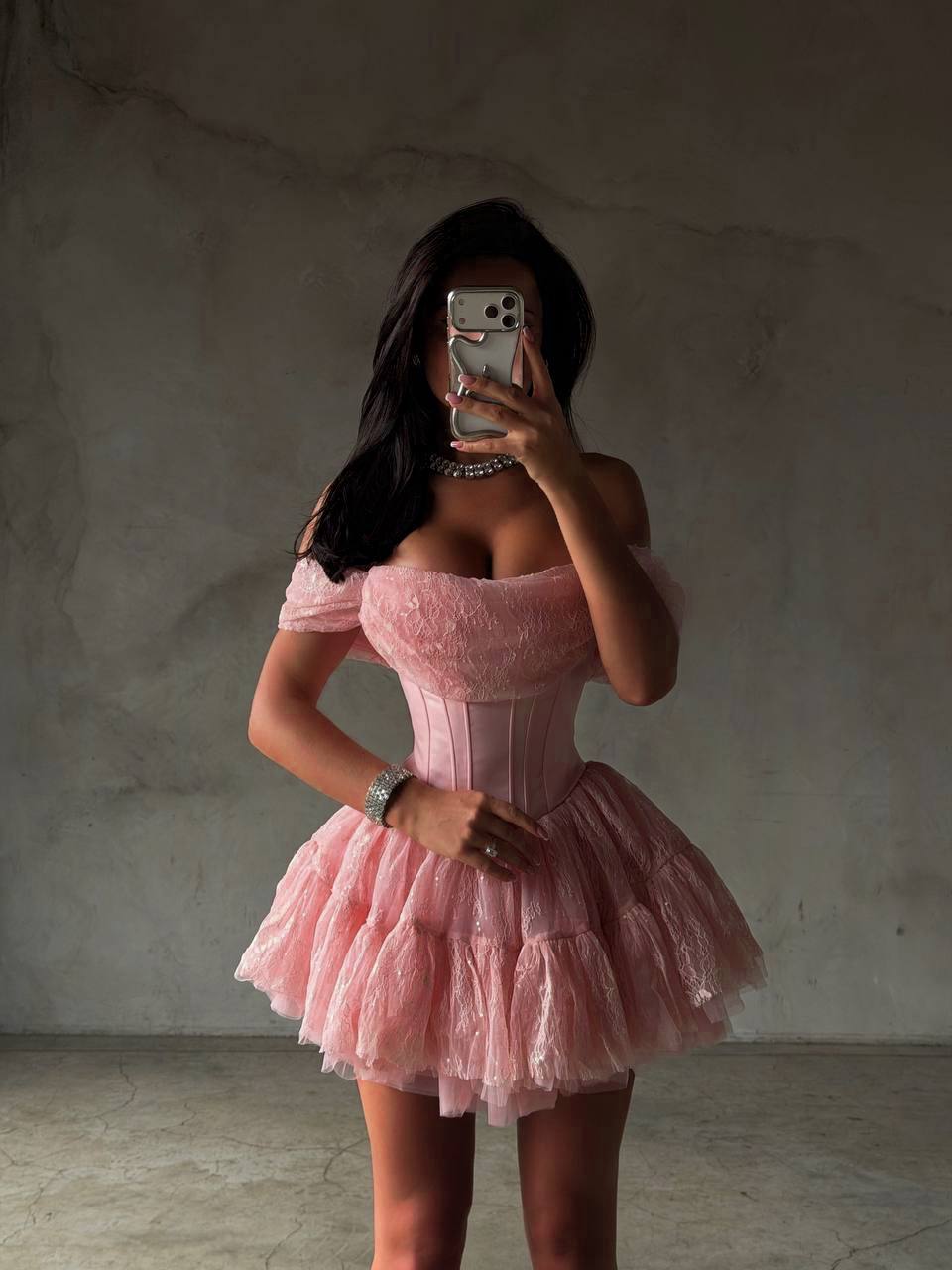 A woman takes a mirror selfie wearing a light pink off-the-shoulder corset dress with a structured waist and a layered ruffled tulle skirt, set against a textured gray concrete wall.