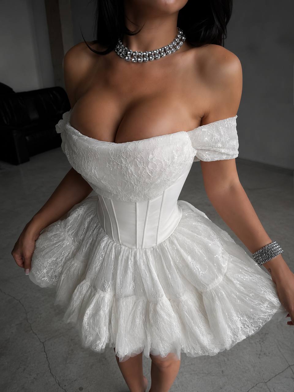 A woman wearing a short off-the-shoulder white lace dress with a structured corset-style bodice and a fluffy ruffled skirt, paired with a rhinestone choker and bracelet.