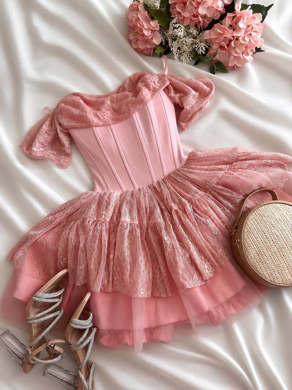 A pink off-the-shoulder corset-style dress with a ruffled tulle skirt, styled in a flat lay on white satin fabric with pink flowers, a woven handbag, and strappy heels.