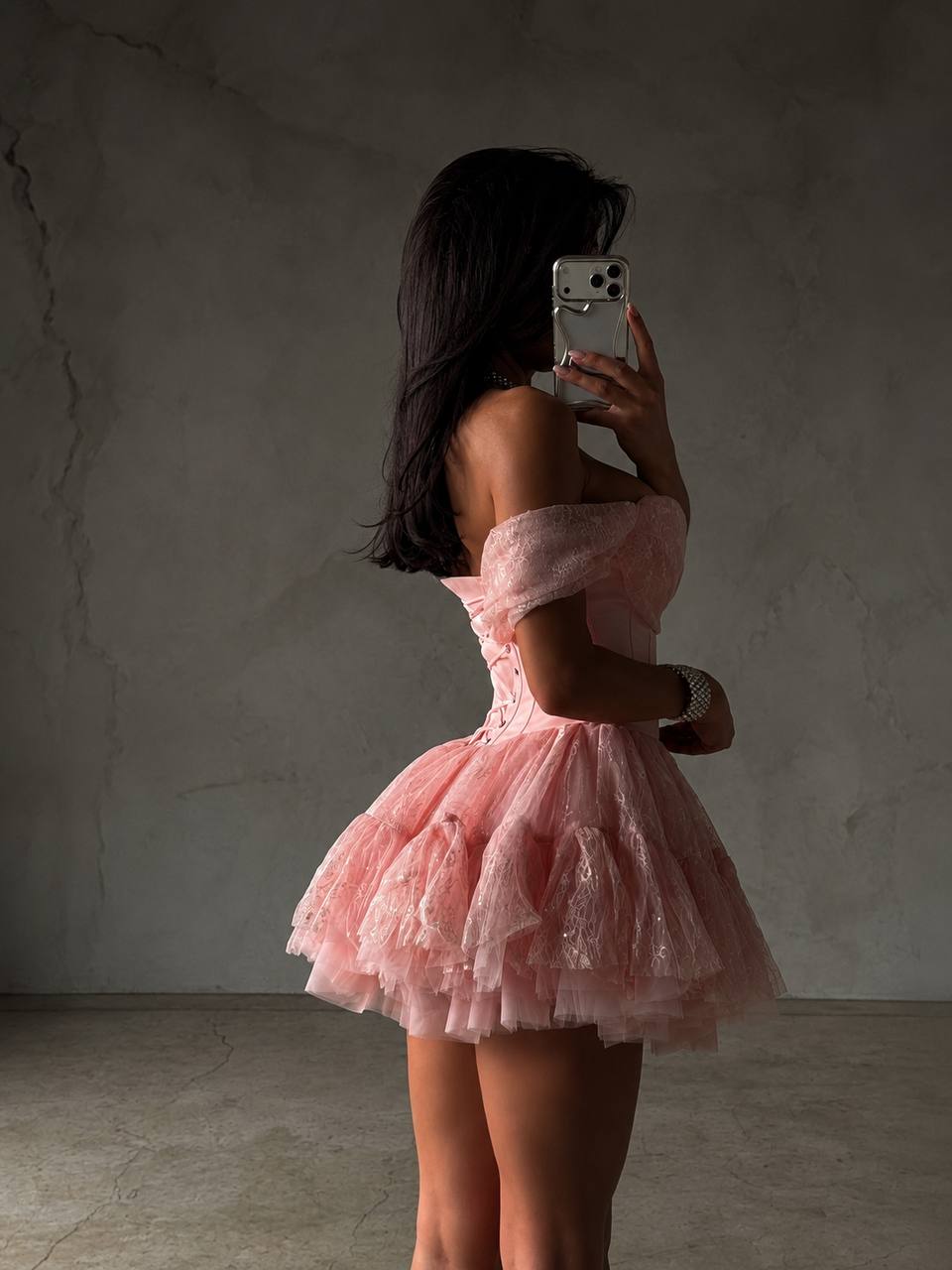 A woman takes a mirror selfie wearing a light pink off-the-shoulder tulle mini dress with a ruffled skirt, standing against a textured gray concrete wall.