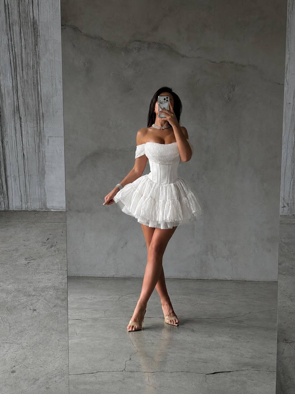 A woman takes a mirror selfie wearing a short white off-the-shoulder tulle dress with a fitted corset-style bodice and a flared layered skirt, standing in a minimalist concrete room.