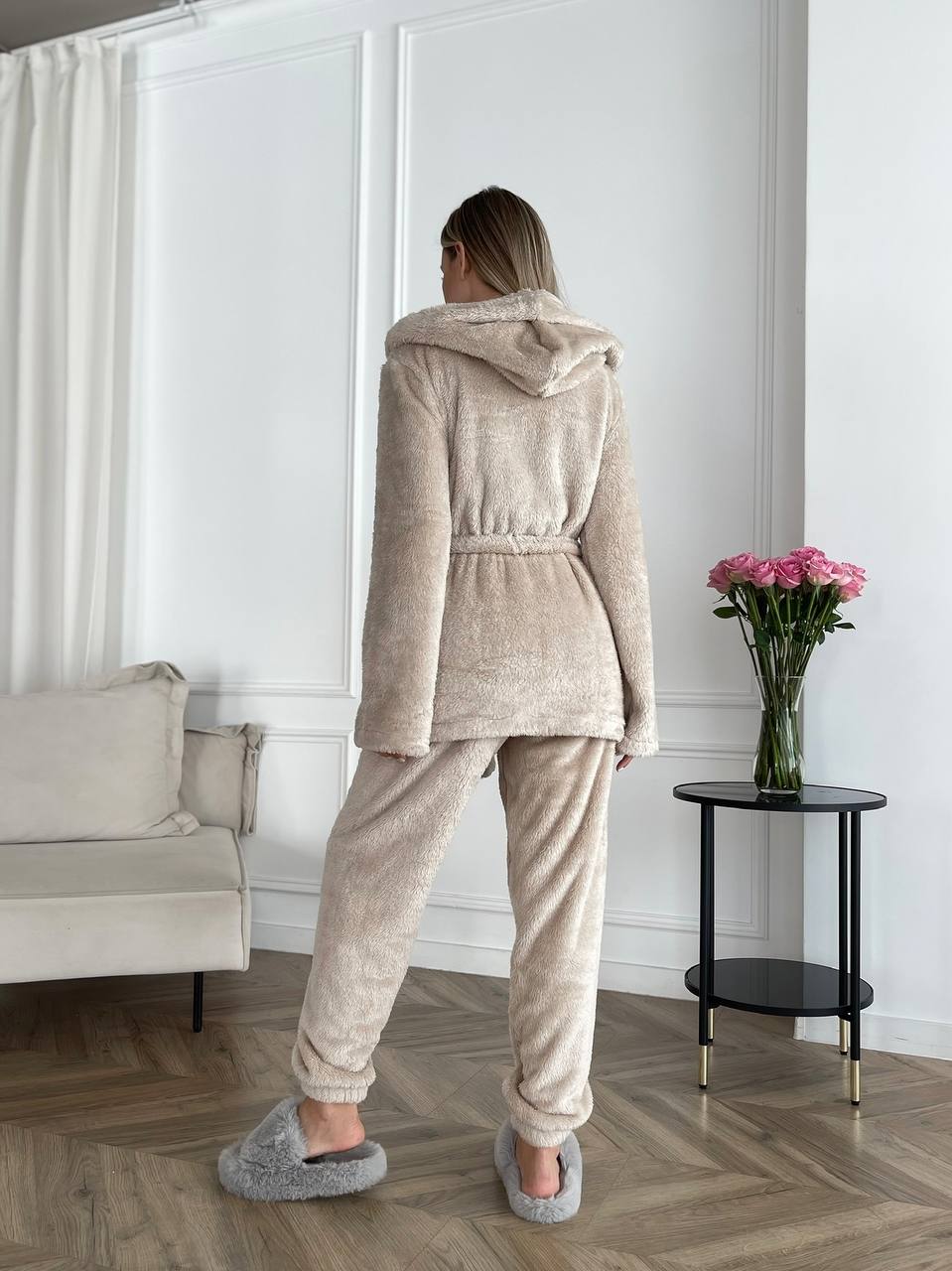 Person wearing a beige robe and slippers in a room with a white wall and a vase of flowers on a table.