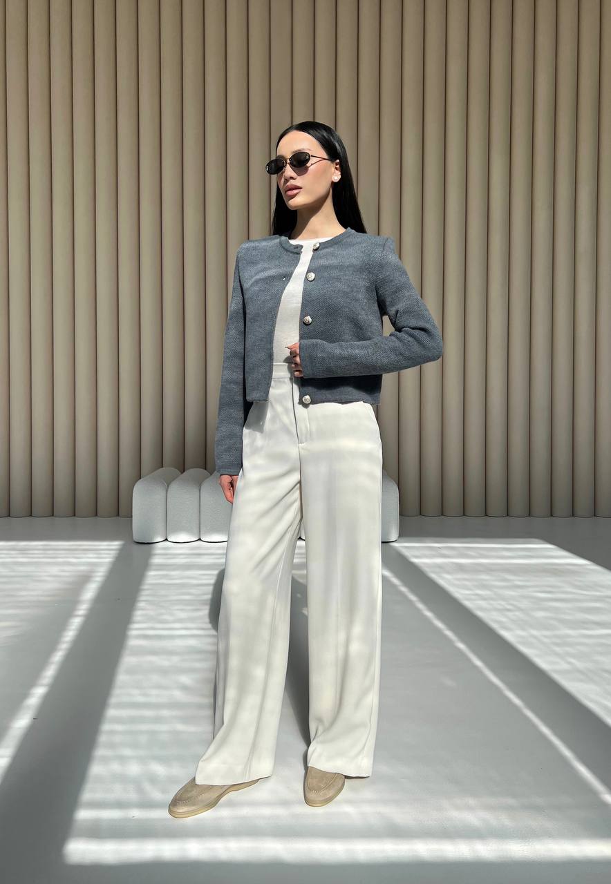 Woman wearing a gray jacket, white top, and white pants against a beige wall.