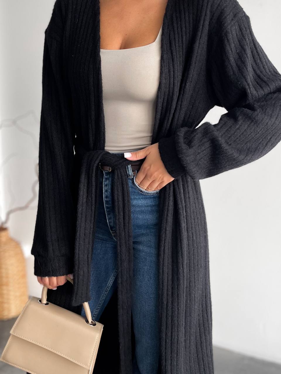 Person wearing a long dark gray cardigan over a beige top and blue jeans, holding a beige handbag.