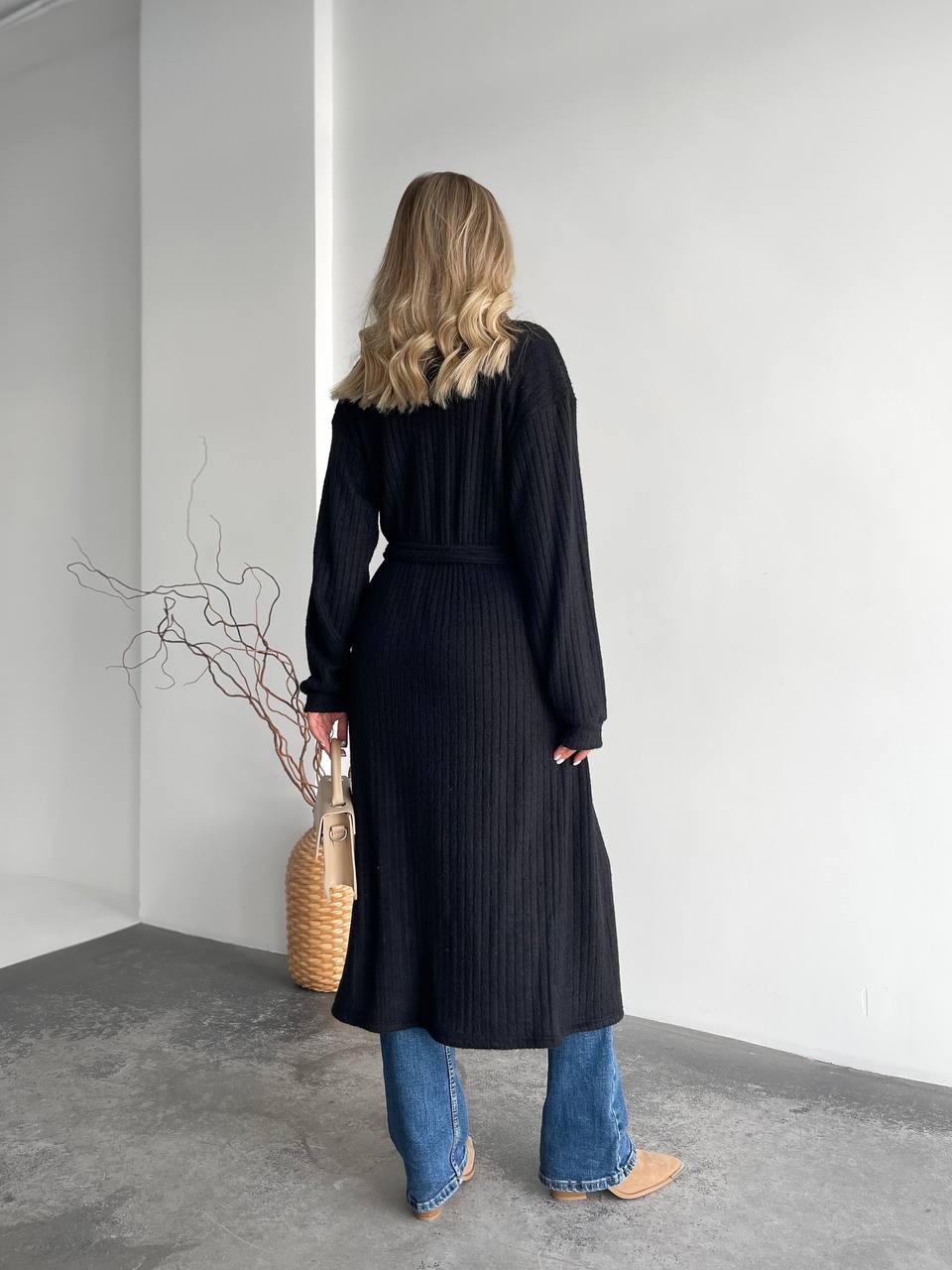 Person wearing a long black coat and blue jeans standing against a white wall.