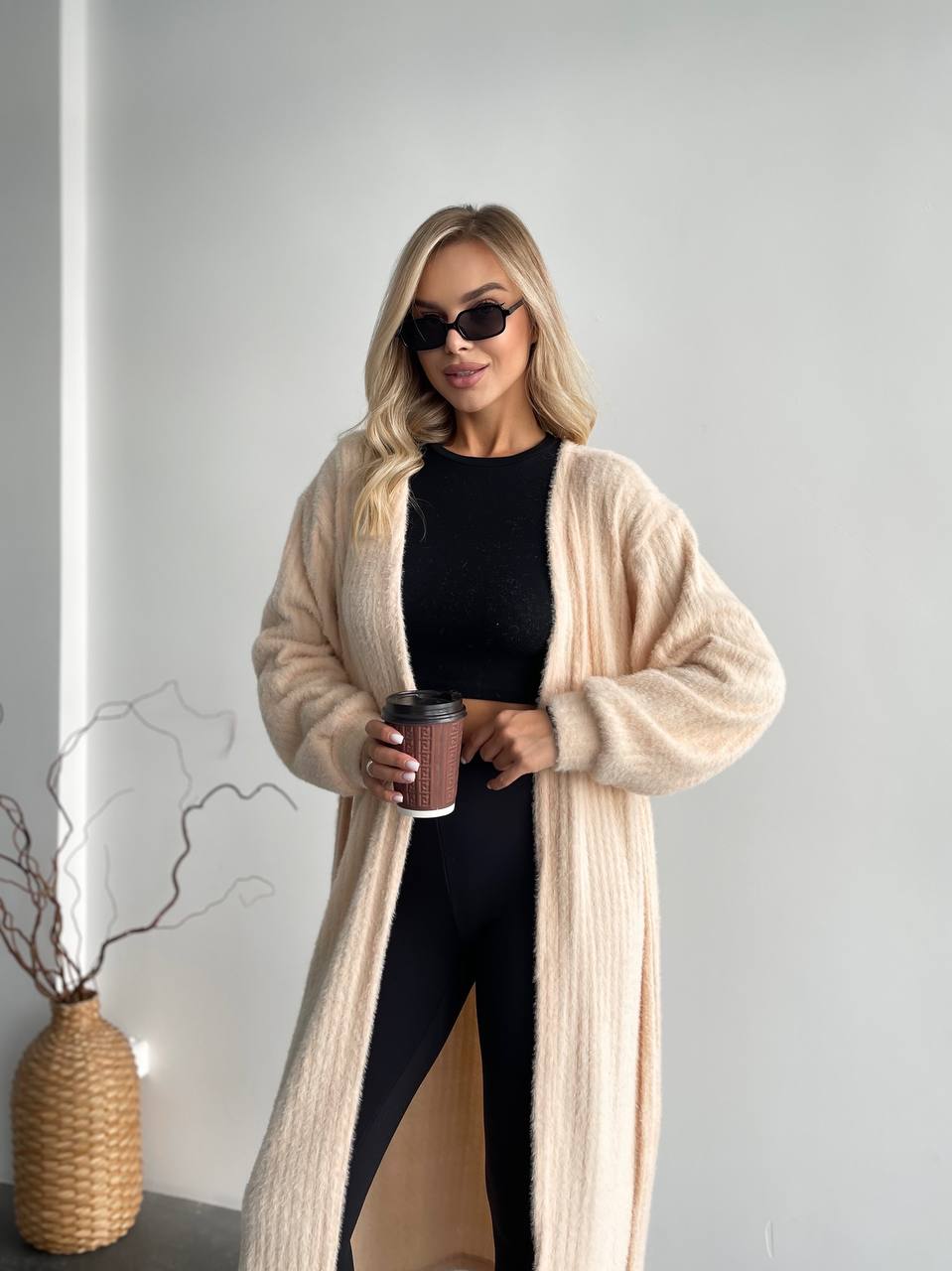 Woman wearing a long beige cardigan and black pants, holding a coffee cup indoors.