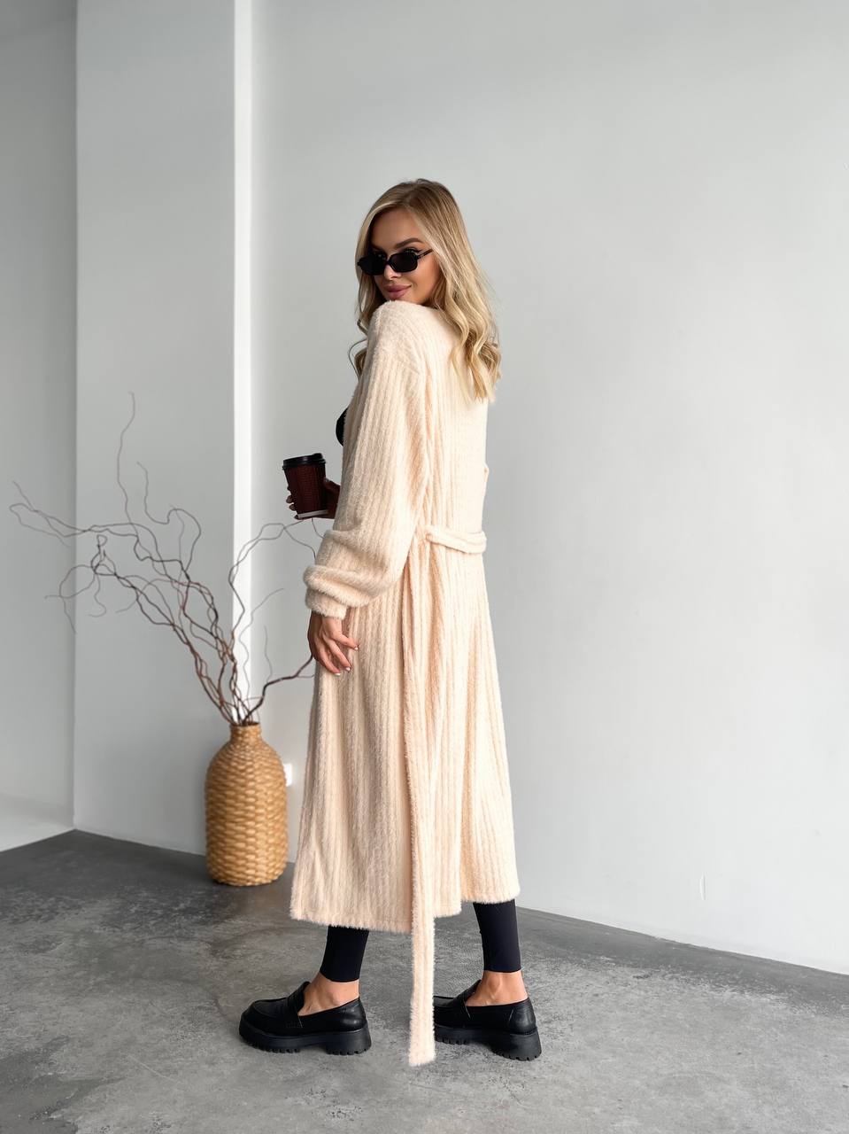 Woman in a long beige cardigan standing in a minimalistic room with a vase and plant.