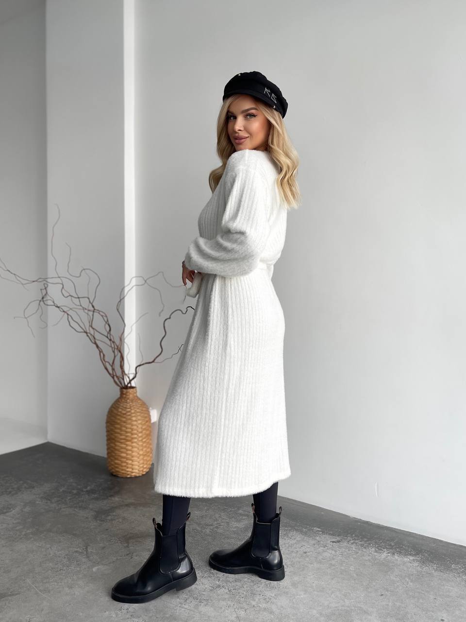 Woman wearing a long white coat and black boots in a minimalistic setting.