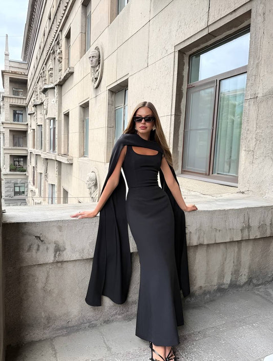 Woman in a black dress with a cape standing against a building.
