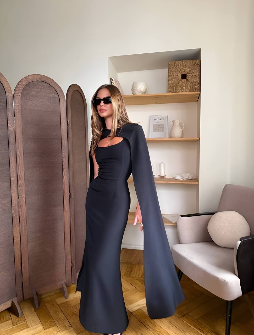 Woman in a long black dress standing in a room with wooden flooring and a shelf in the background.