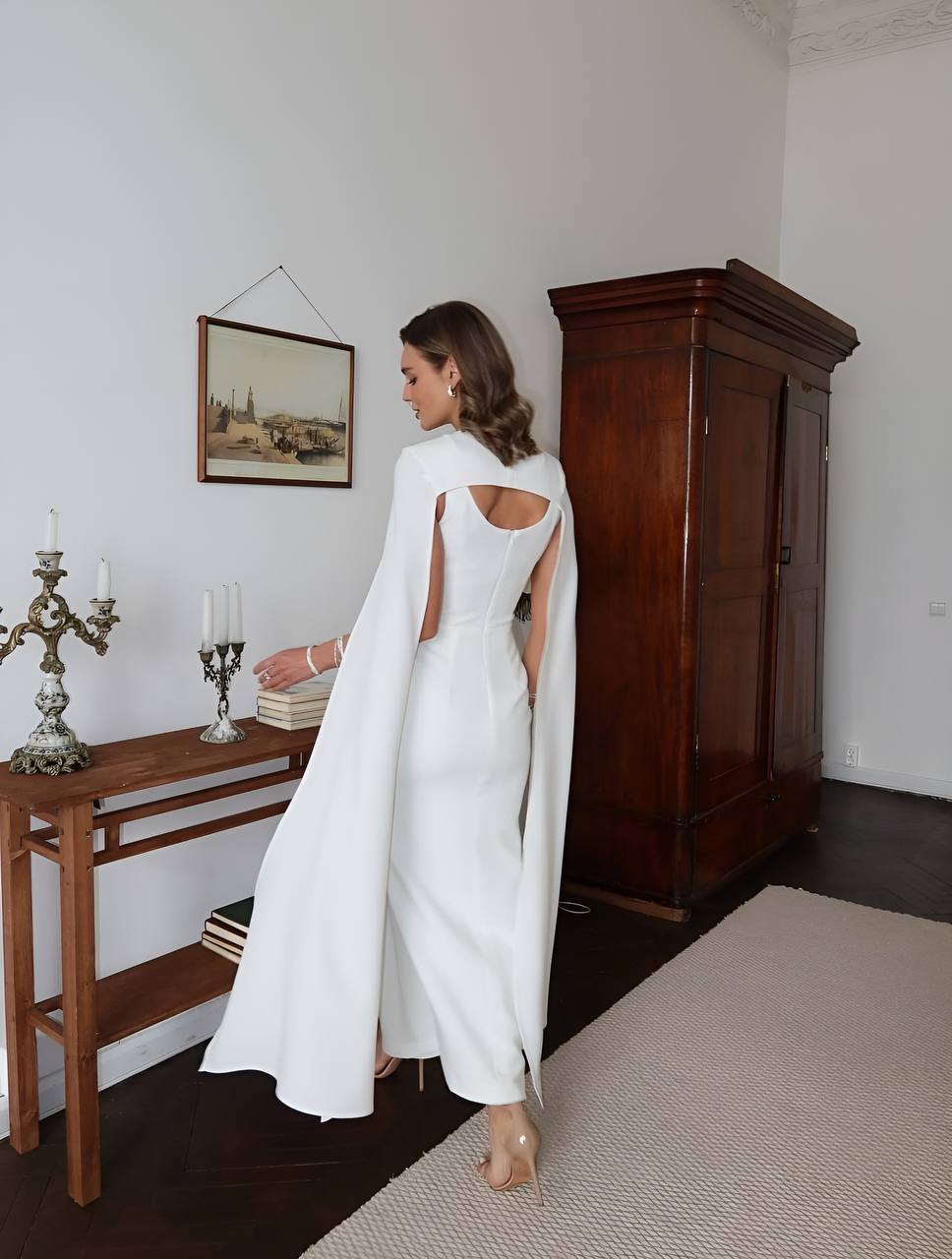 Woman in a white dress with cut-out back standing in a room with wooden furniture and a painting on the wall.