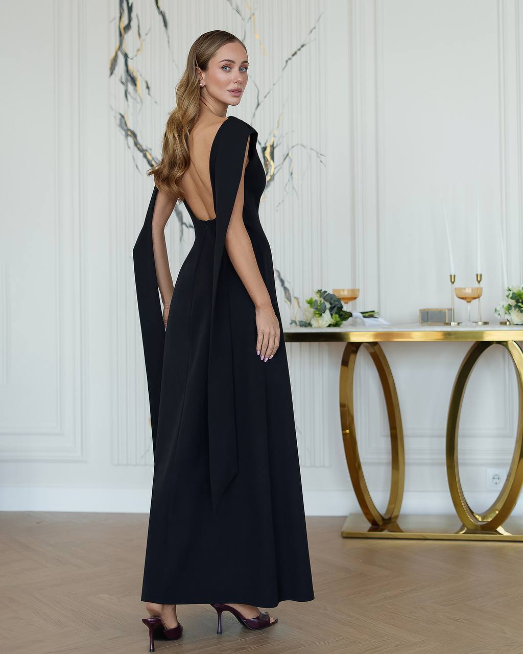 Woman in a black evening gown standing in a stylish interior setting with a gold console table.