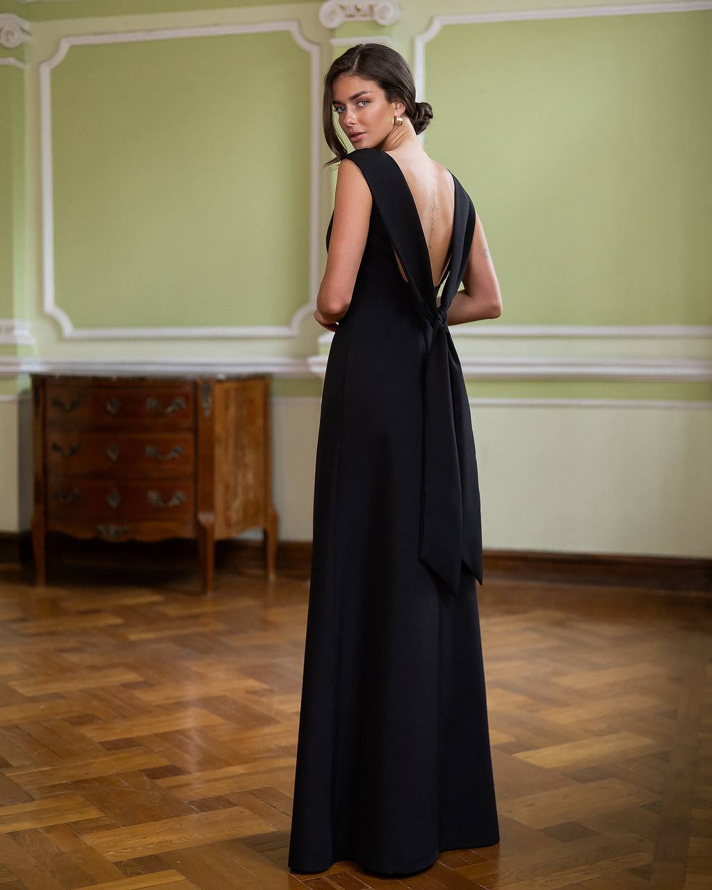 Woman in a black evening gown standing in an elegant room with wooden floor and green walls.