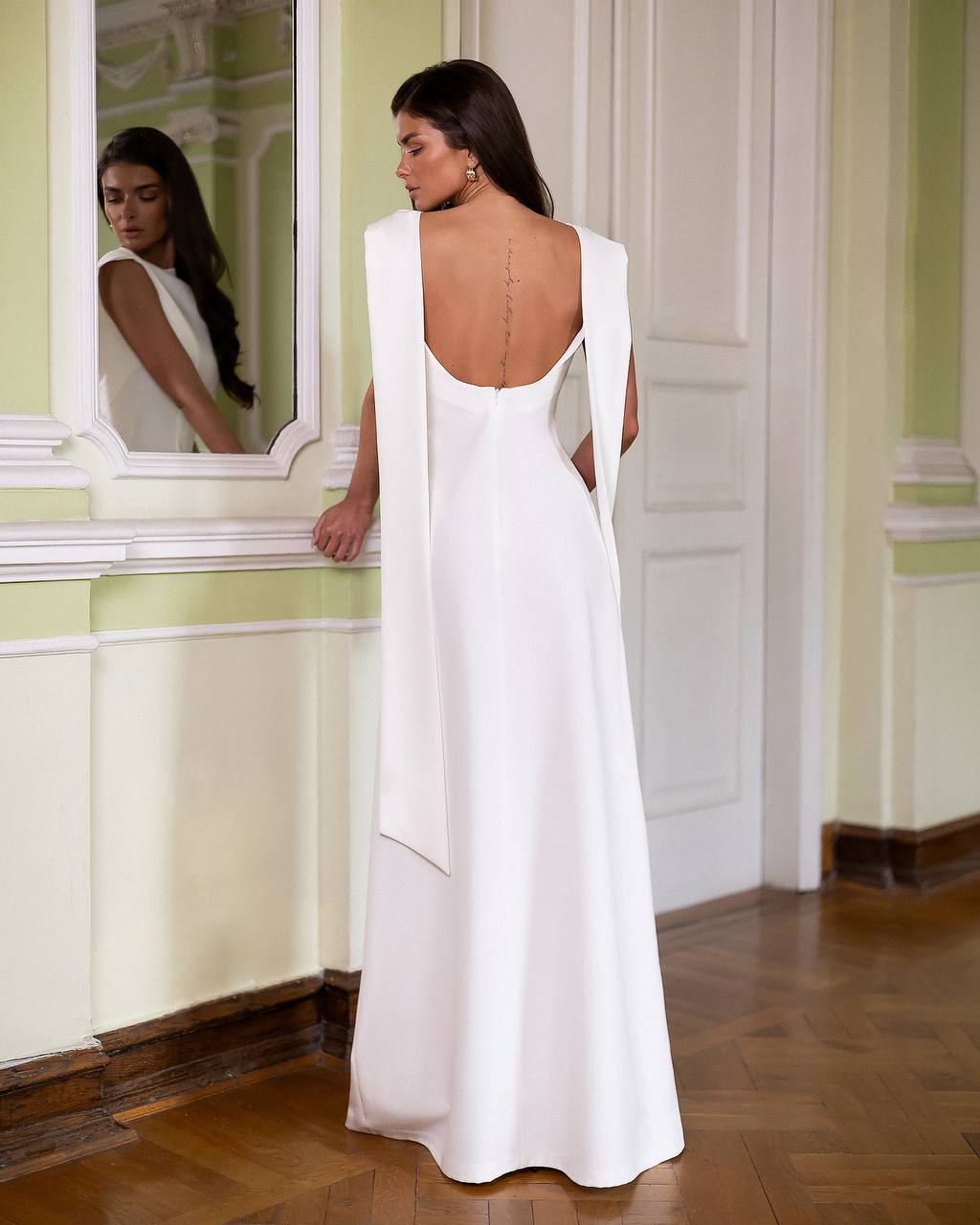 Woman in a white evening gown standing in a room with a mirror.