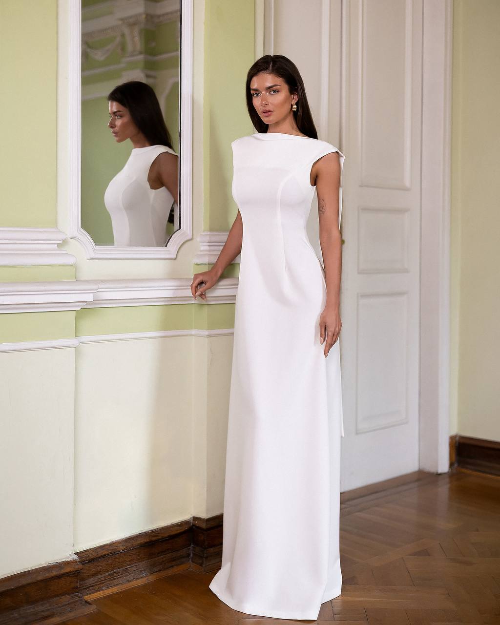 Woman in a white dress standing in a room with a mirror reflecting her.