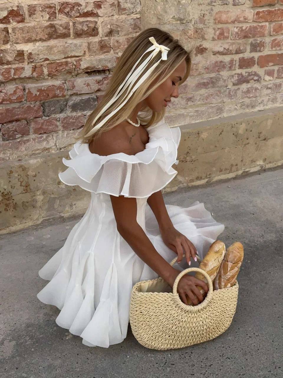 Woman in a white dress holding a basket with bread against a brick wall.