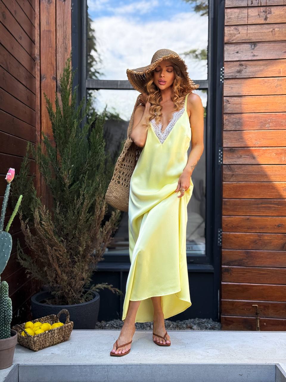 Woman in a yellow dress and straw hat standing in front of a wooden building with plants around.