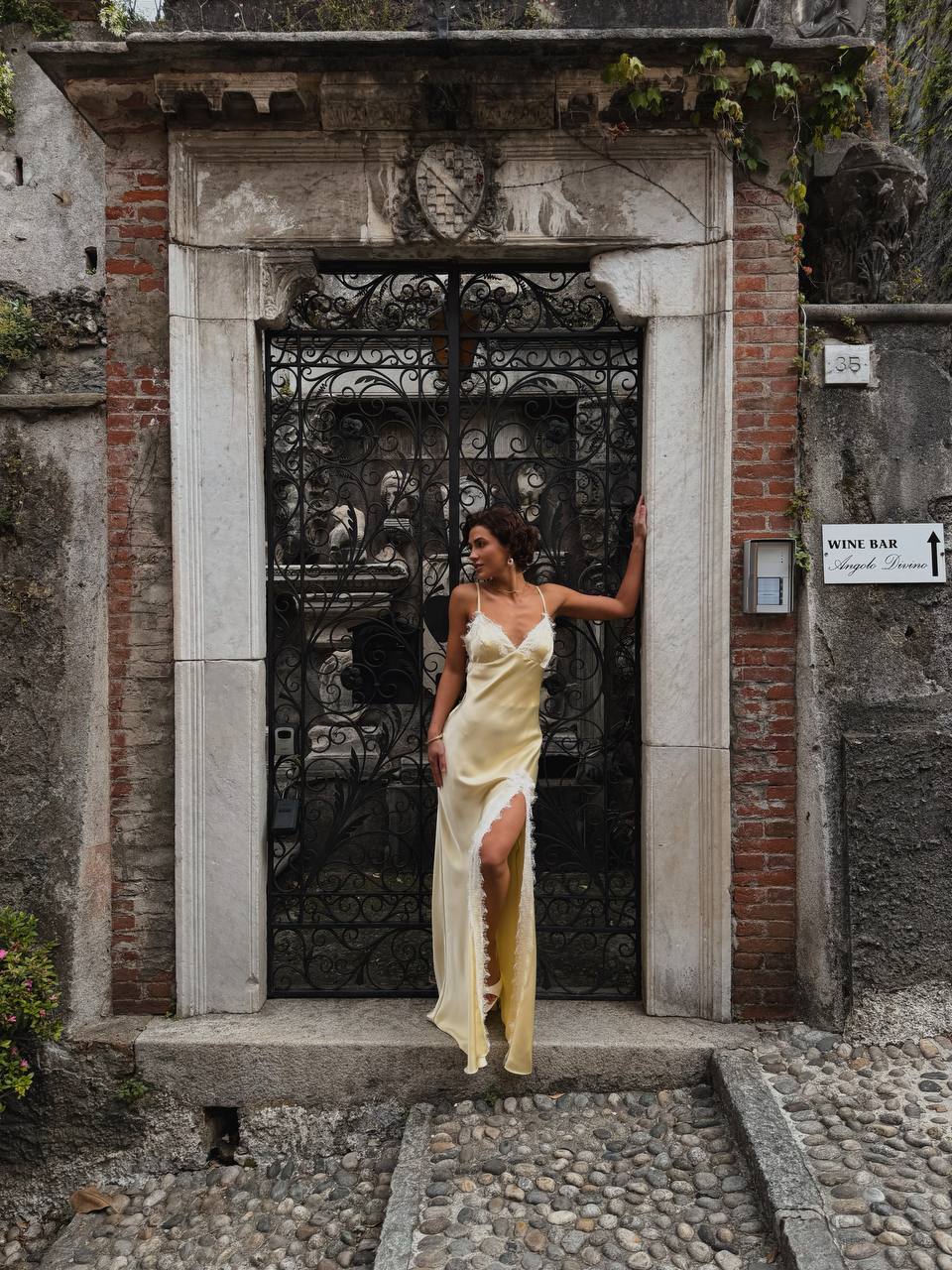A woman wearing a pale yellow satin slip dress with a thigh-high slit stands in front of an old stone and iron gate doorway on a cobblestone street.