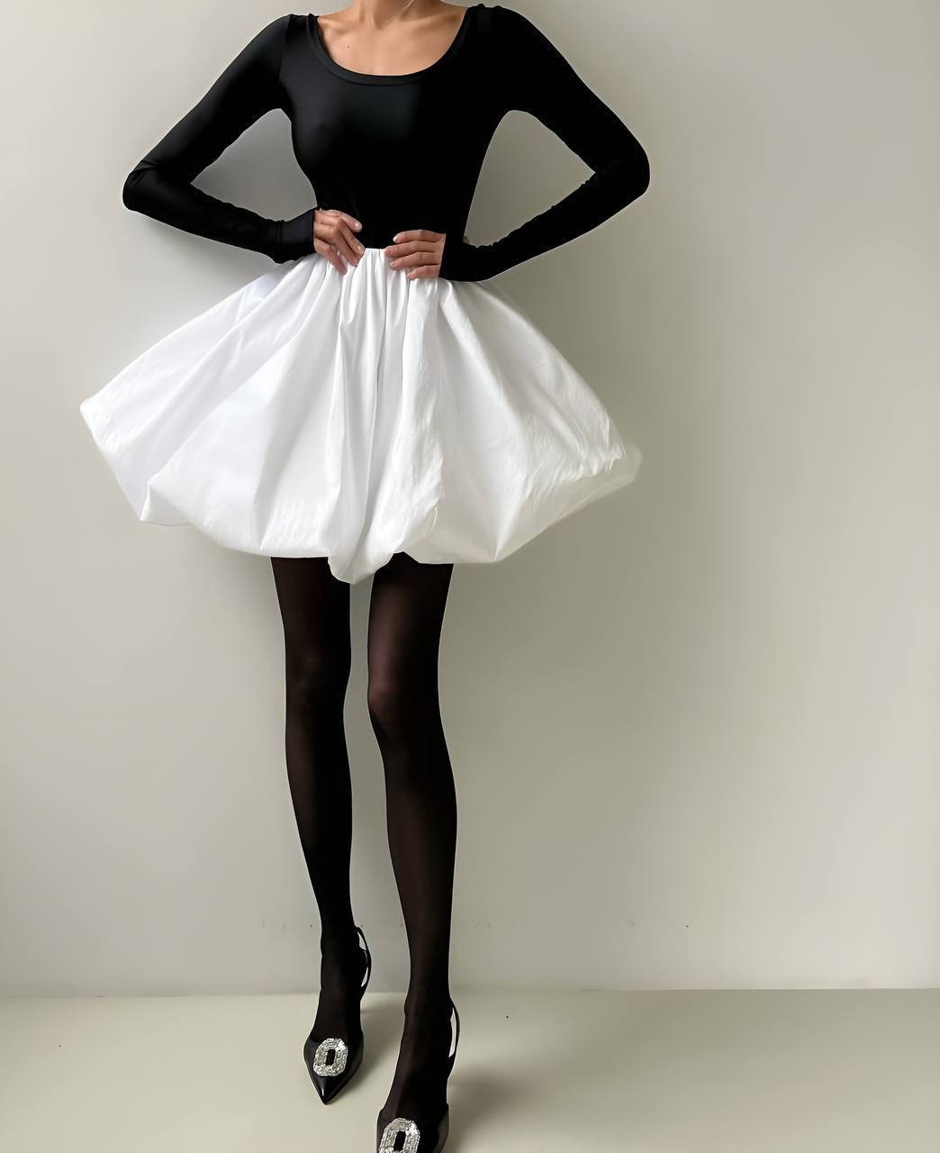 Person wearing a black long-sleeve top and white skirt with black tights and shoes on a plain background