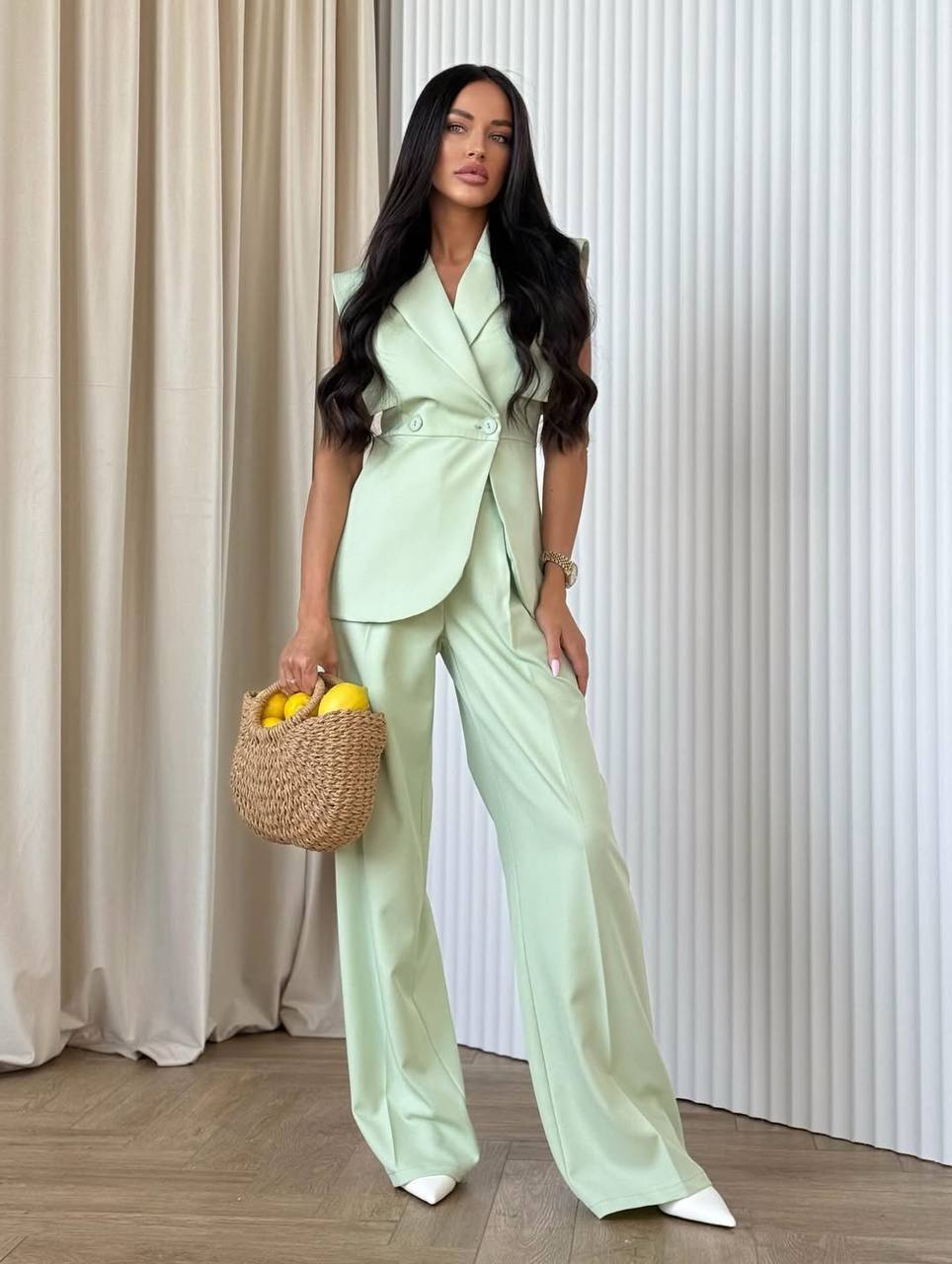 Woman in a light green suit holding a woven basket with lemons against a neutral background