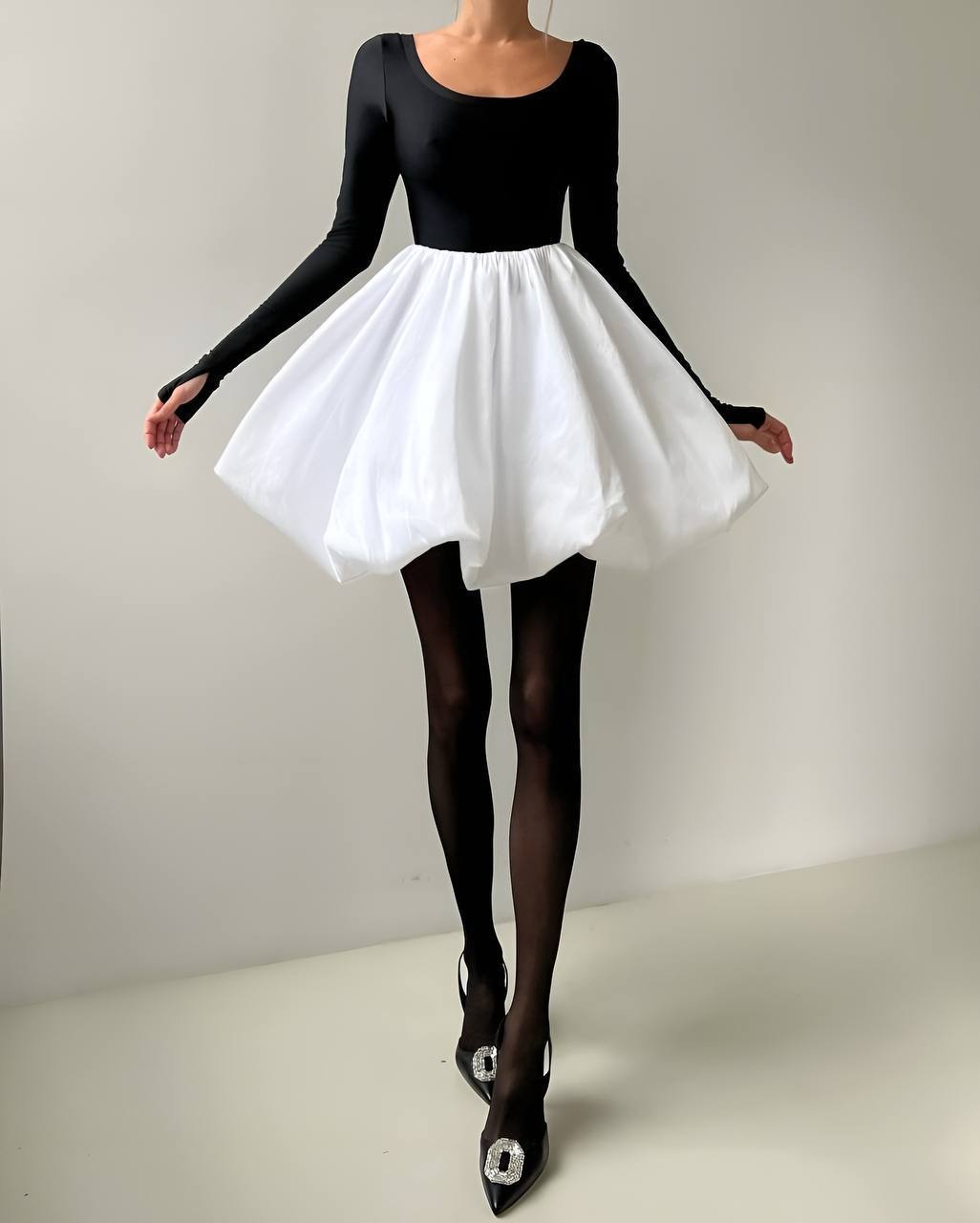 Person wearing a black long-sleeve top and white skirt with black tights and shoes on a plain background