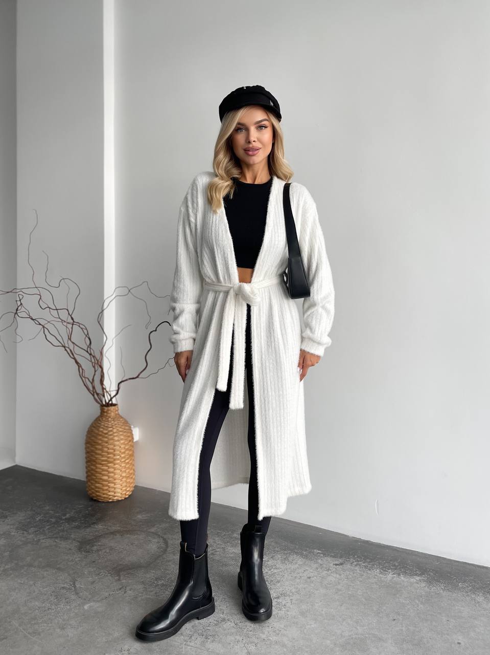 Woman wearing a white coat, black top, and black pants in a minimalistic setting.