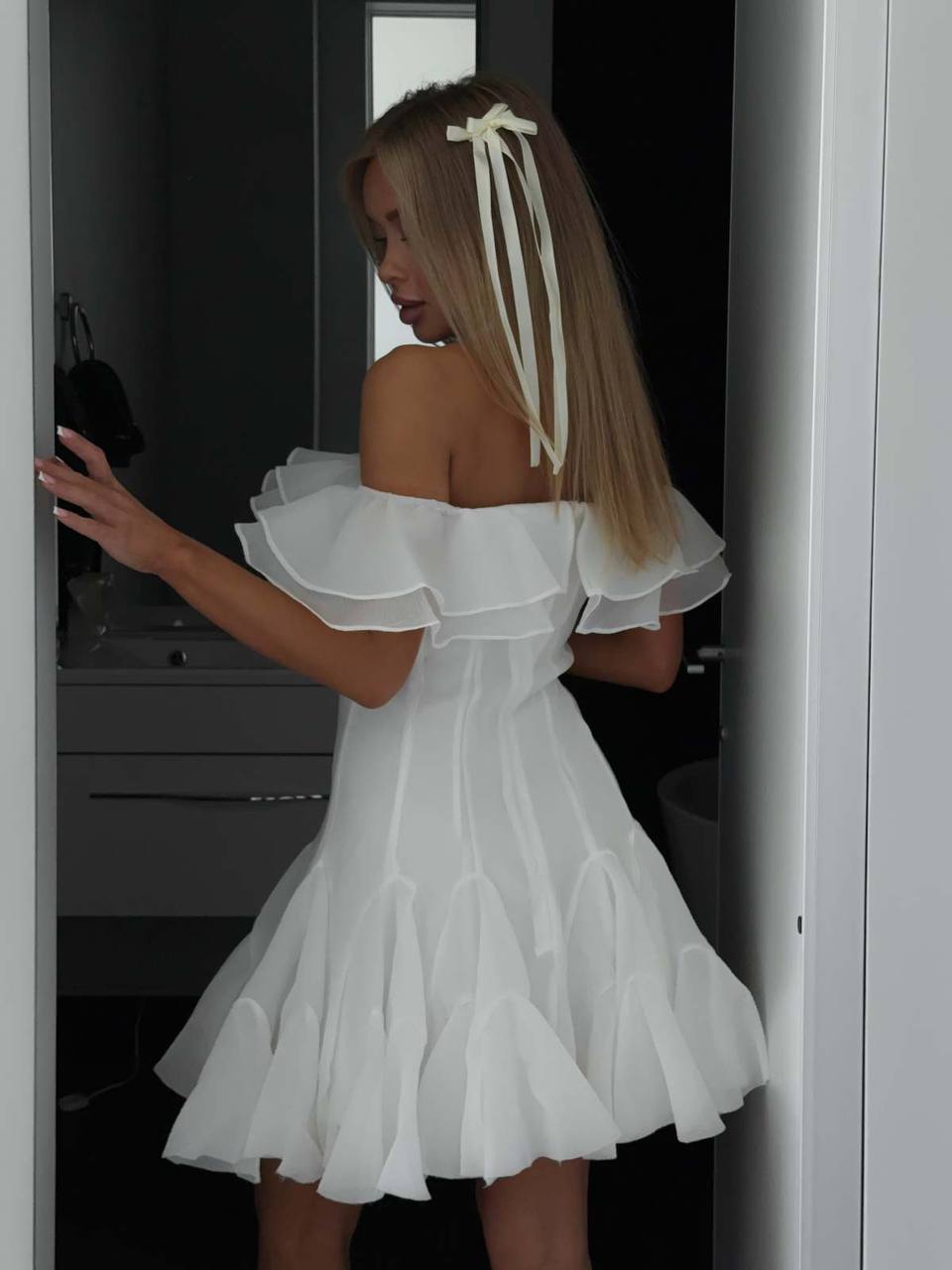 Woman wearing a white off-shoulder dress with ruffled details, standing in a doorway.