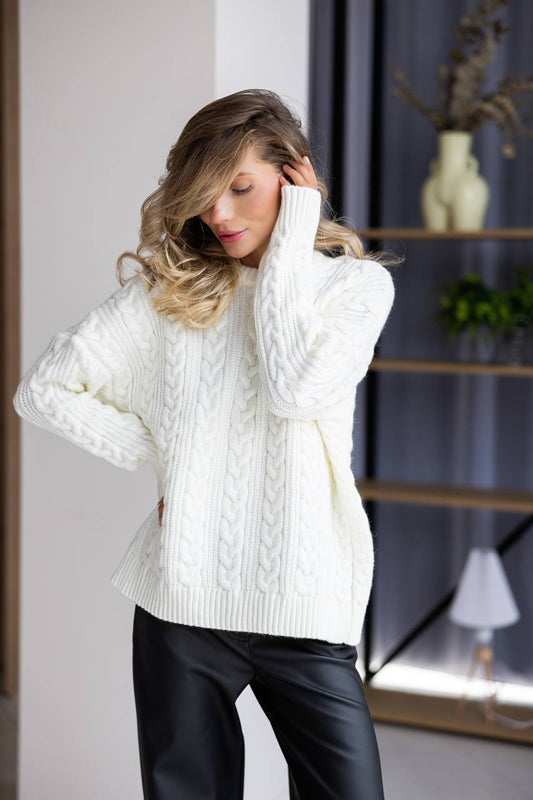 Woman wearing a white cable knit sweater and black pants indoors.
