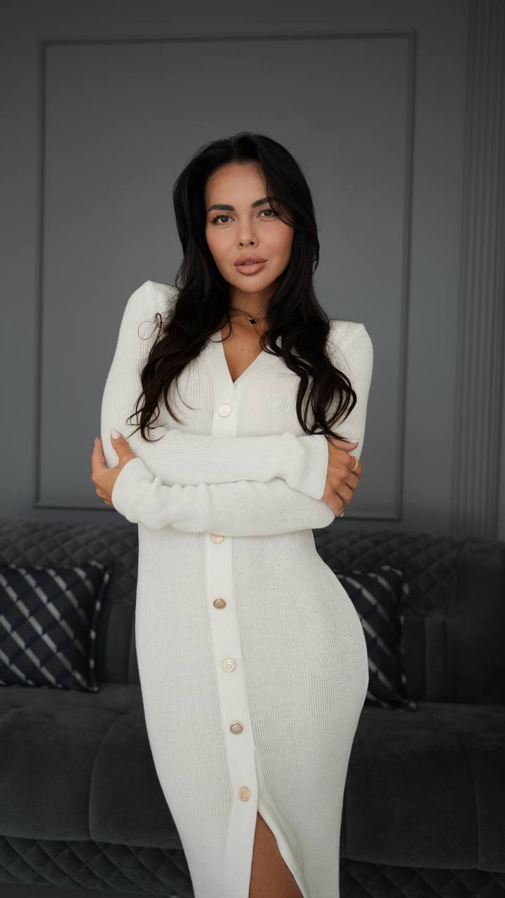 Woman wearing a white dress with a button detail against a gray wall.