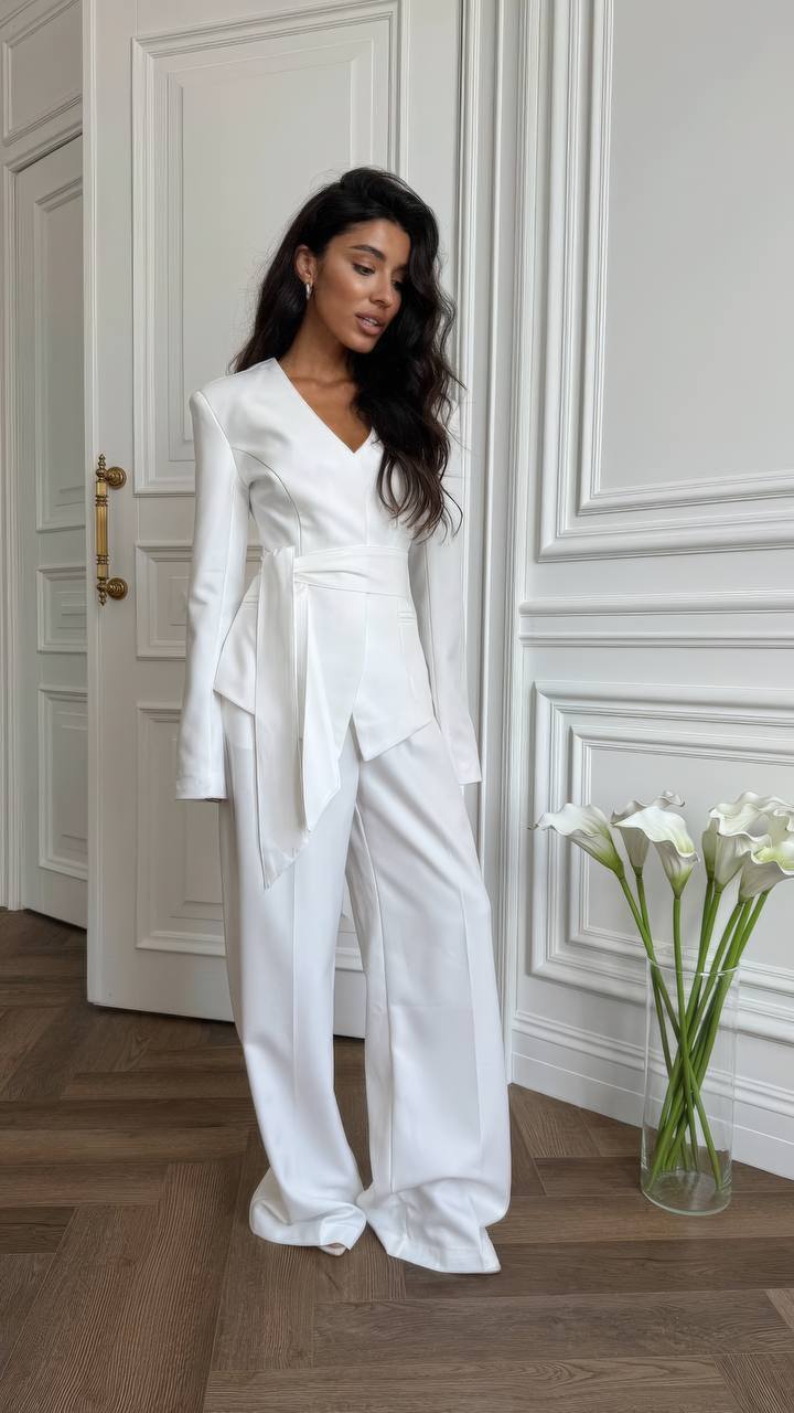 Woman in a white outfit standing in a room with a vase of flowers.