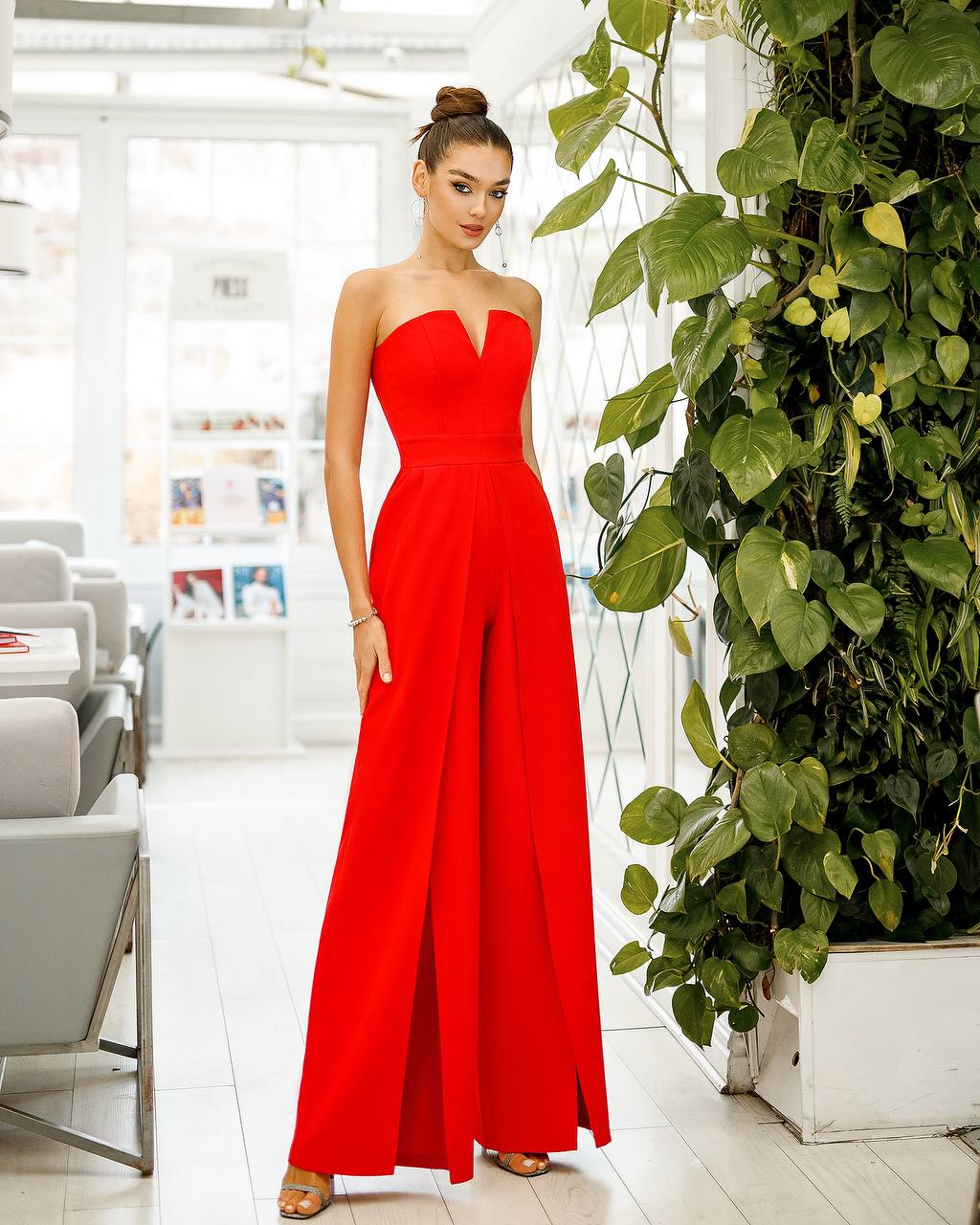 Woman in a red strapless jumpsuit standing in a modern indoor setting with plants.