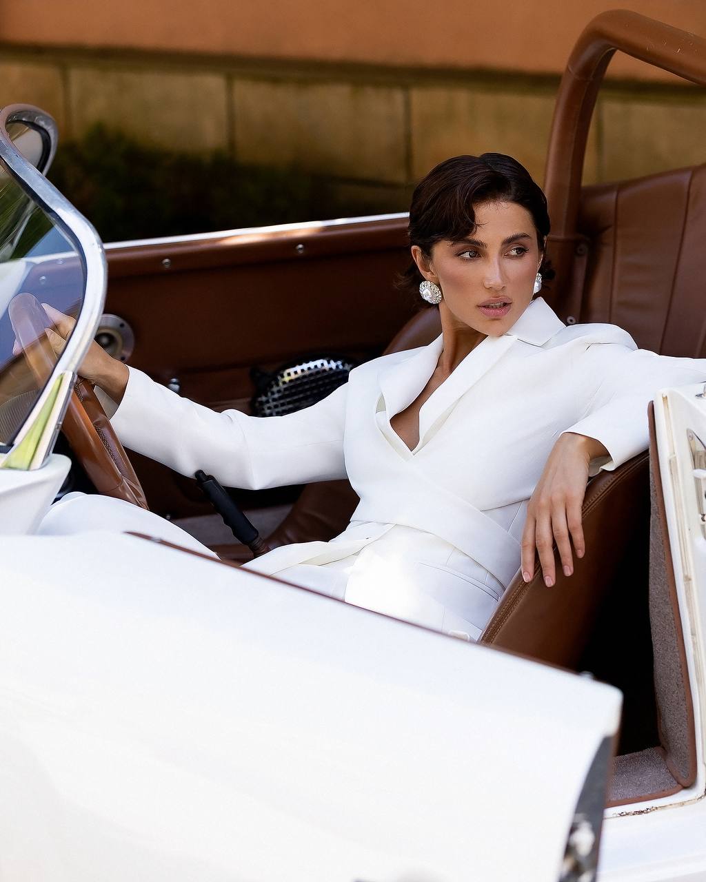 A woman wearing a white tailored blazer and matching trousers sits in a brown leather convertible car, with the car's white exterior visible in the foreground.