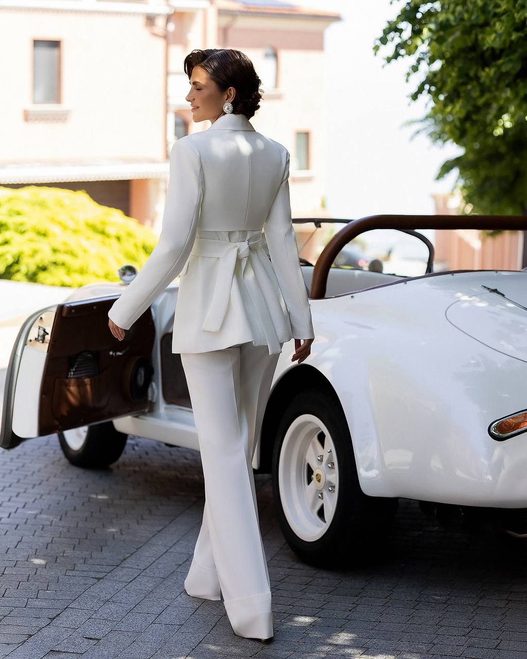 A woman wearing a white belted blazer and matching wide-leg trousers stands beside a white vintage convertible car outdoors.