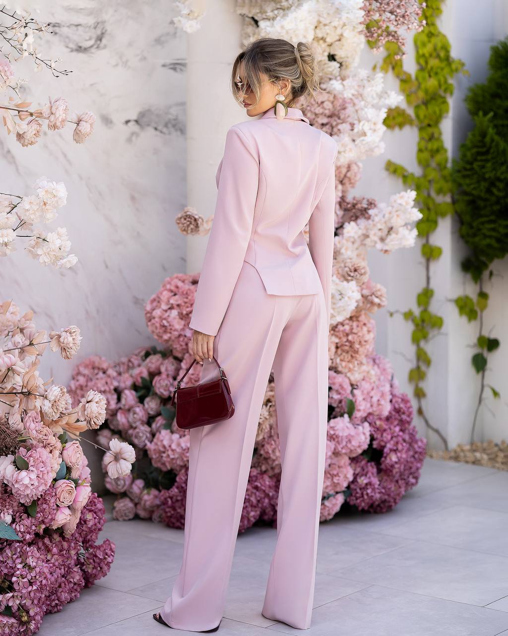 A woman stands with her back to the camera wearing a light pink tailored suit with a structured jacket and wide-leg trousers, holding a small dark burgundy handbag. She is posed in front of a decorative wall covered in pink and white flowers and green vines.