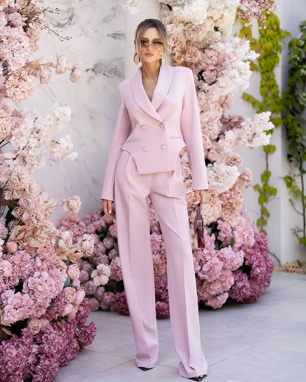 A woman stands outdoors in front of a floral wall wearing a light pink tailored suit with a double-breasted blazer and matching trousers. She is holding a small dark handbag.
