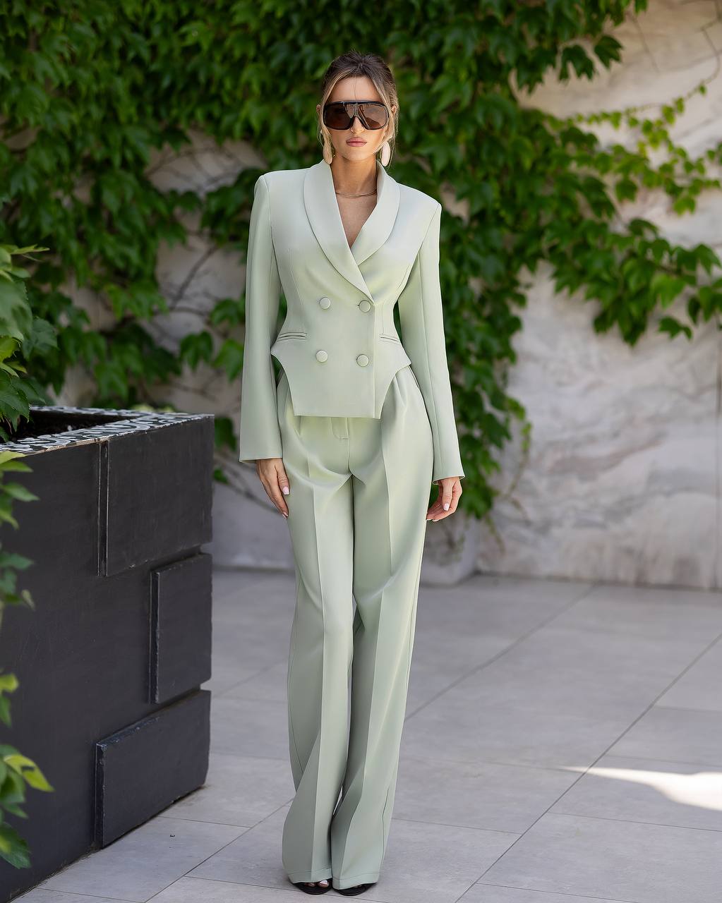 A woman models a light sage green double-breasted blazer and matching wide-leg trousers set, styled with dark sunglasses, standing outdoors against a stone wall with green ivy.