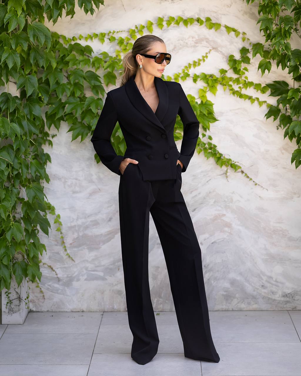 A woman modeling a black double-breasted blazer and matching wide-leg trousers, styled with black sunglasses, standing against a textured white wall with green ivy.