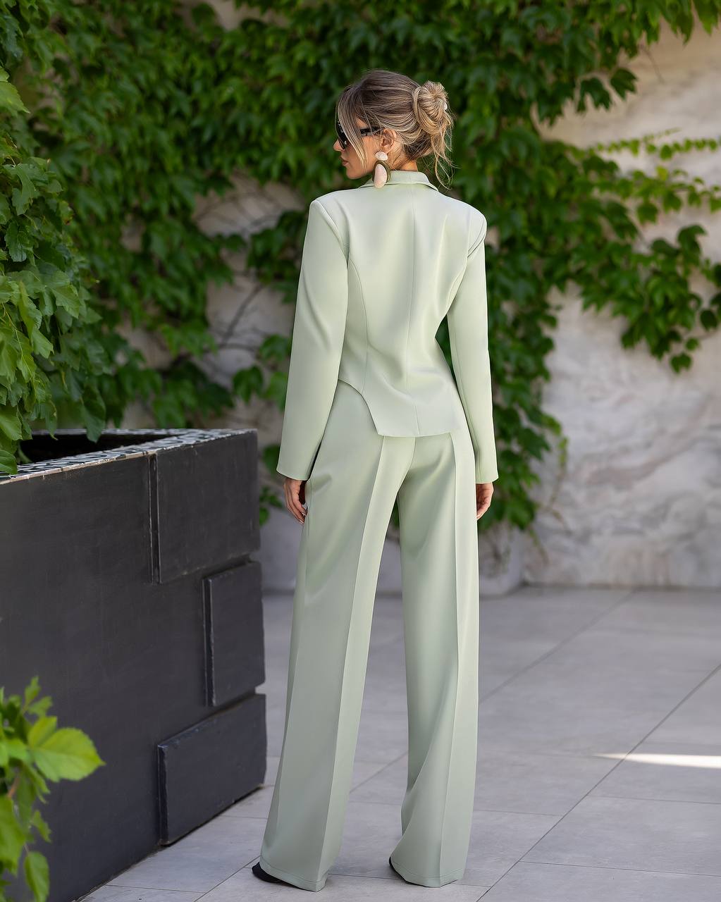 A woman stands outdoors facing away from the camera, wearing a light sage green tailored suit with a structured jacket and wide-leg trousers. The setting features a stone wall covered in green ivy, a dark gray planter, and light gray paving stones.