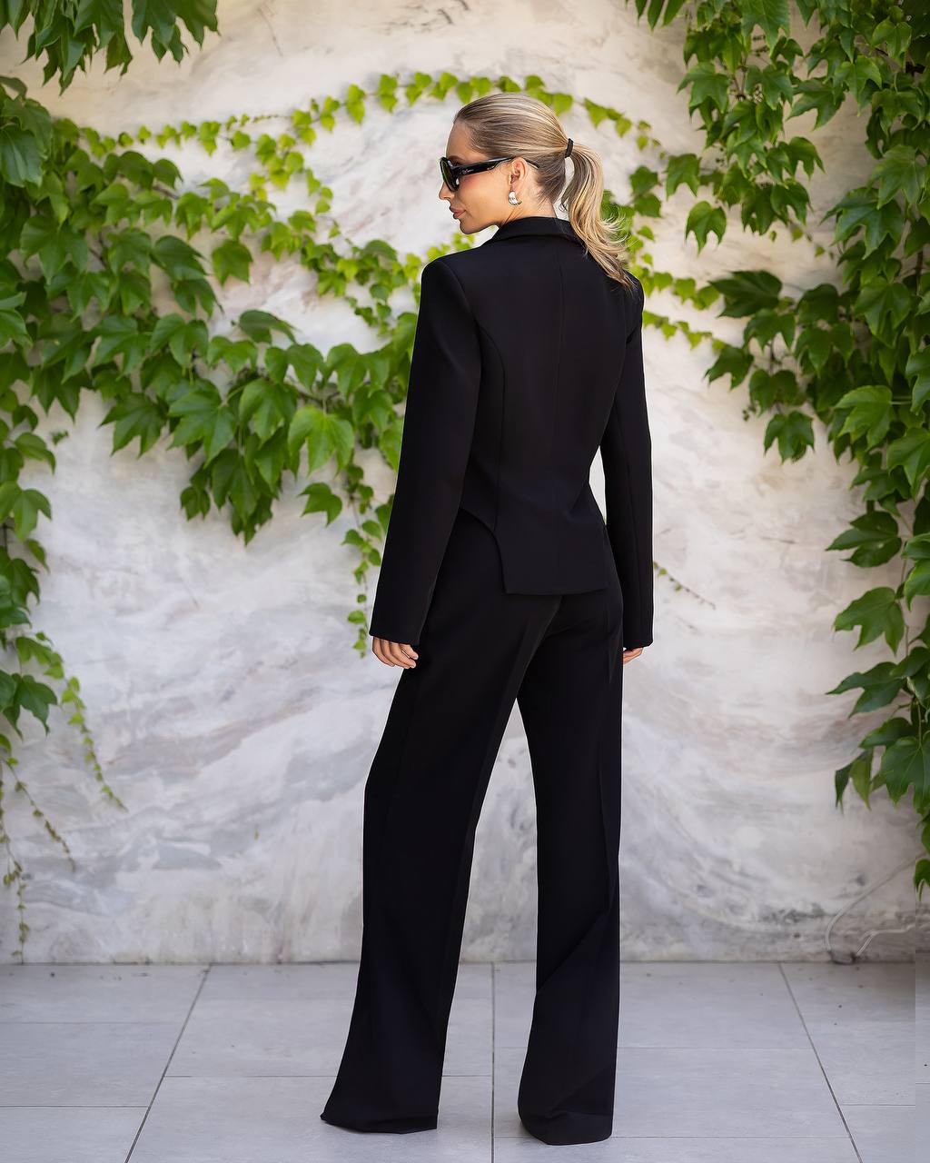 A woman wearing a black tailored pantsuit with a structured blazer and wide-leg trousers, photographed from the back against a light stone wall with green ivy.
