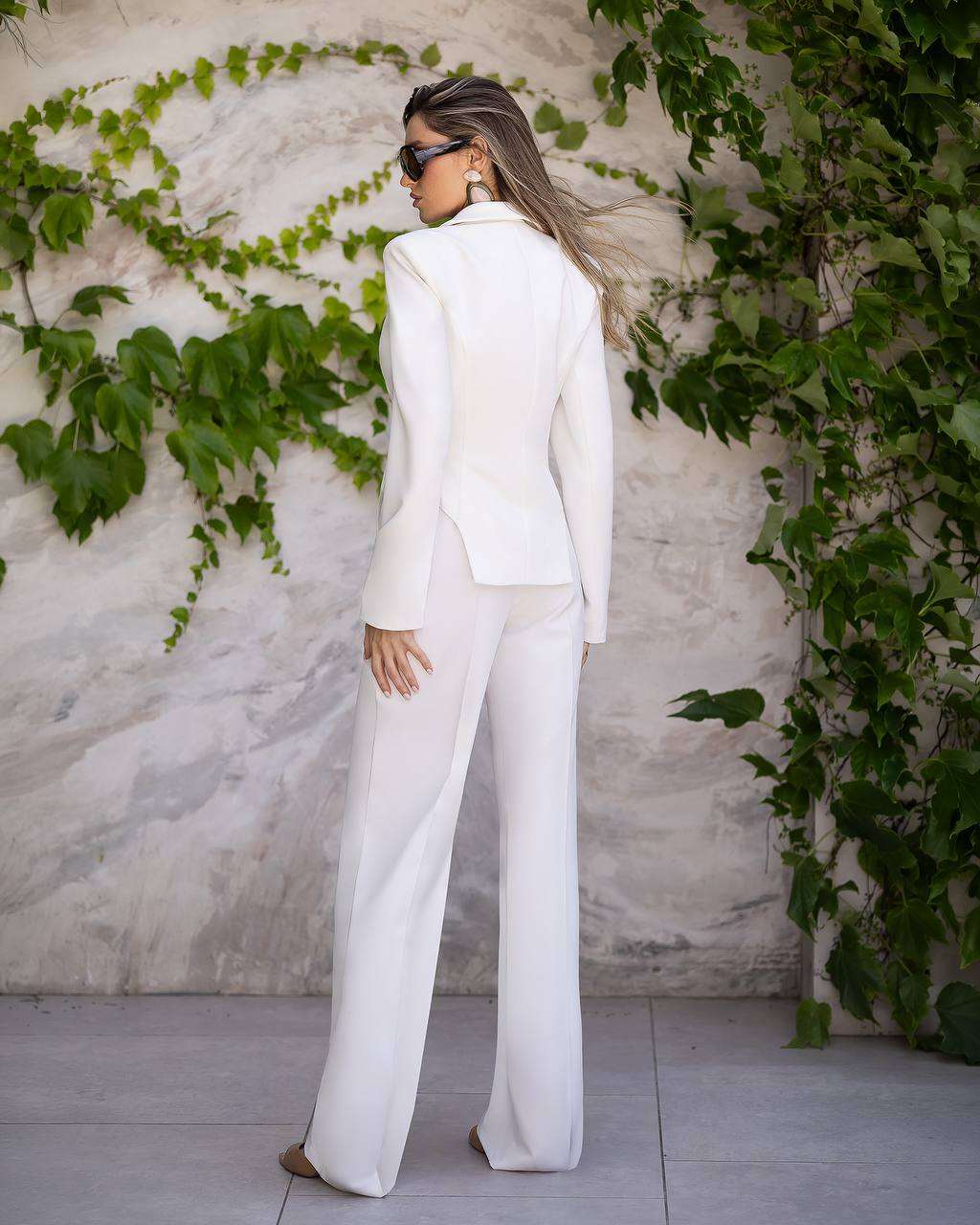 A woman stands with her back to the camera wearing a tailored white blazer and matching wide-leg trousers, posed against a textured light wall with green ivy climbing up it.