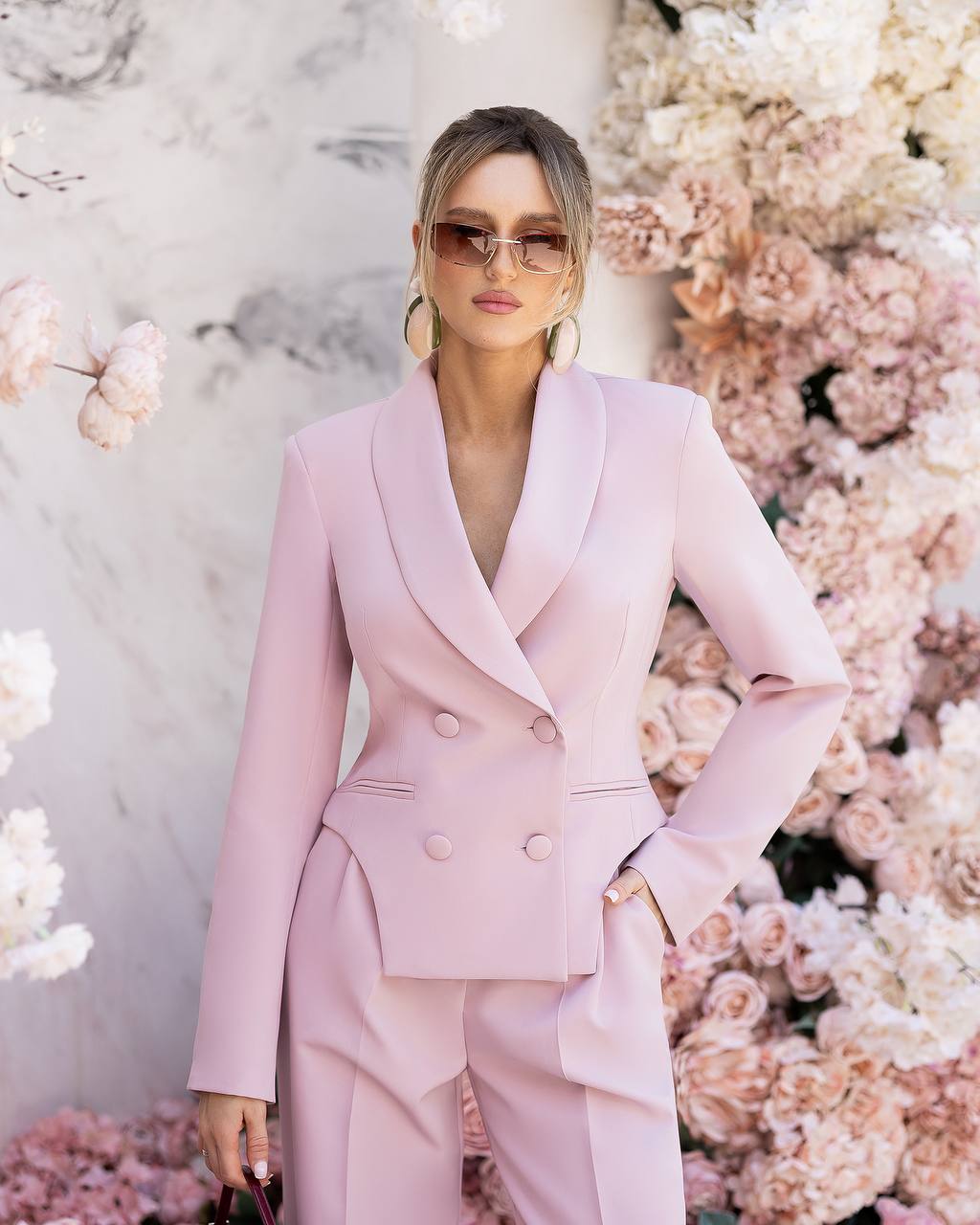A woman models a light pink double-breasted blazer and matching trousers set, styled with gold hoop earrings and sunglasses, standing in front of a floral backdrop.