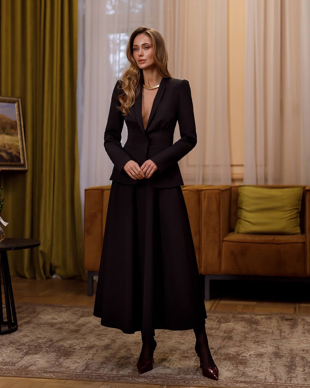 Woman in a black dress standing in an elegant room with green curtains and a brown sofa.