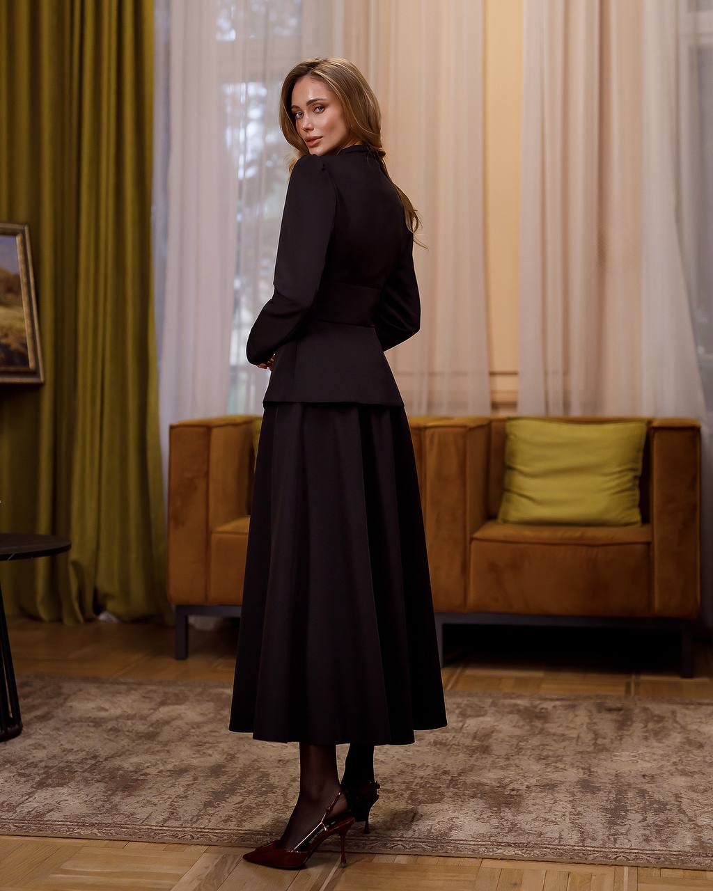 Woman in a black outfit standing in a room with curtains and a couch.